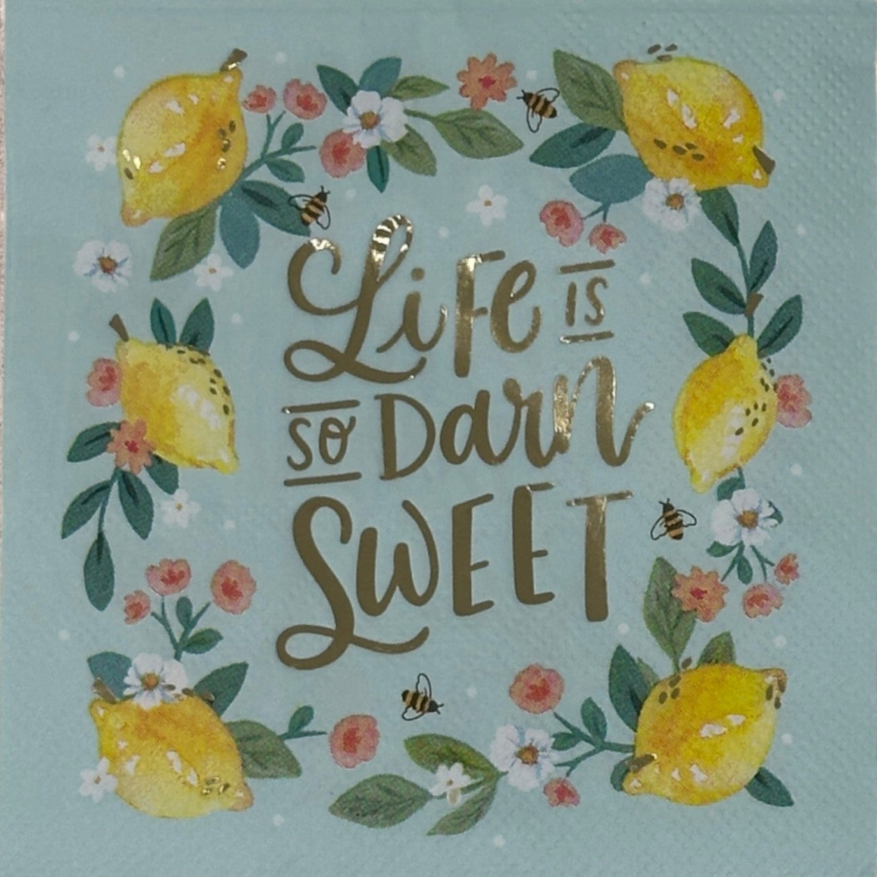 A paper napkin with a blue background featuring yellow lemons, flowers, and the text 'Life is so Darn Sweet' in a gold foil-like script.
