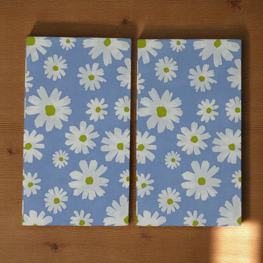 Two guest-size paper napkins with white daisy flowers and green centers on a light blue background, displayed on a wooden surface.
