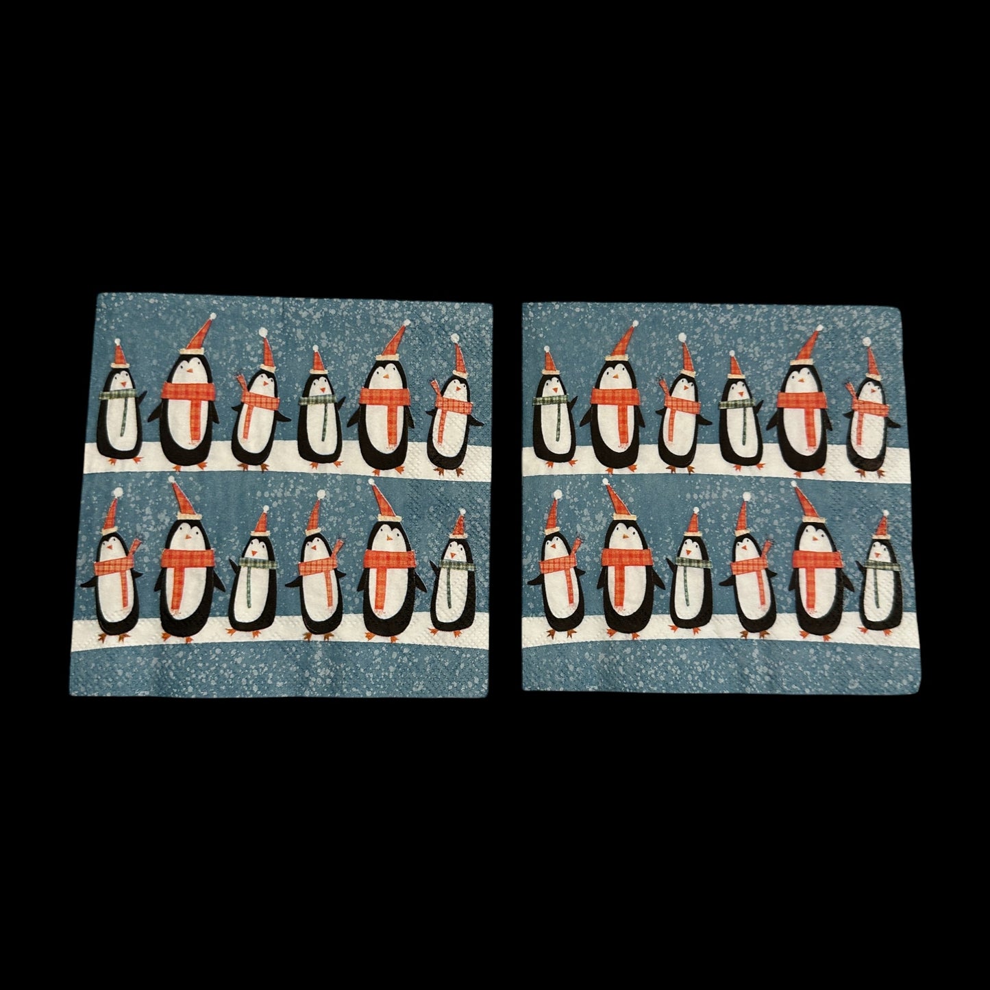 Two Cocktail size decoupage napkin featuring rows of cute penguins wearing scarves and Santa hats on a snowy blue background for holiday crafts and decor.