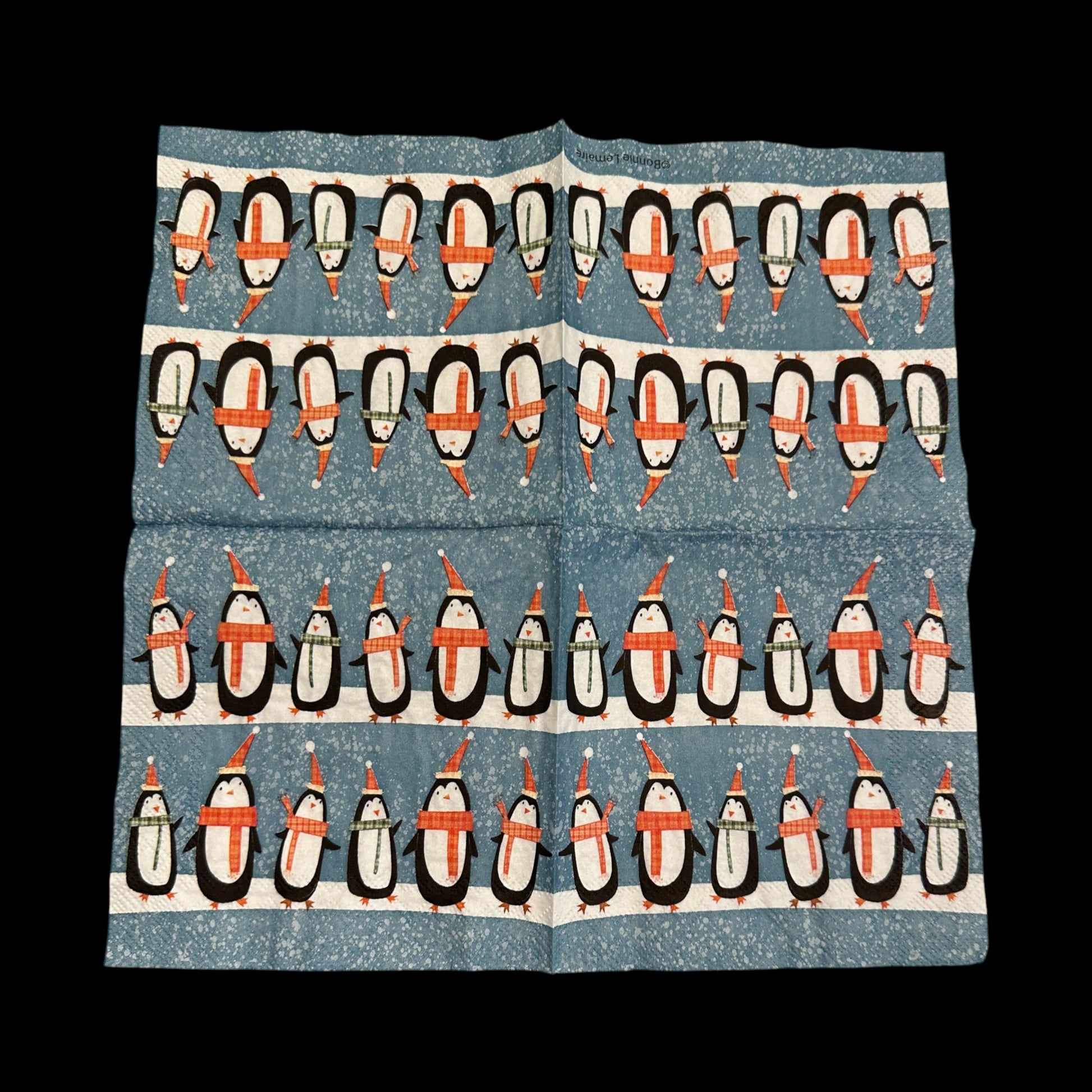 Open unfolded Cocktail size decoupage napkin featuring rows of cute penguins wearing scarves and Santa hats on a snowy blue background for holiday crafts and decor.