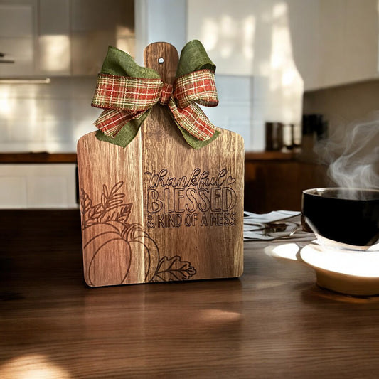 Wood Cutting Board Carved with a Pumpkin and says thankful blessed and kind of a mess.  green and fall colored plaid ribbons are tied around the top