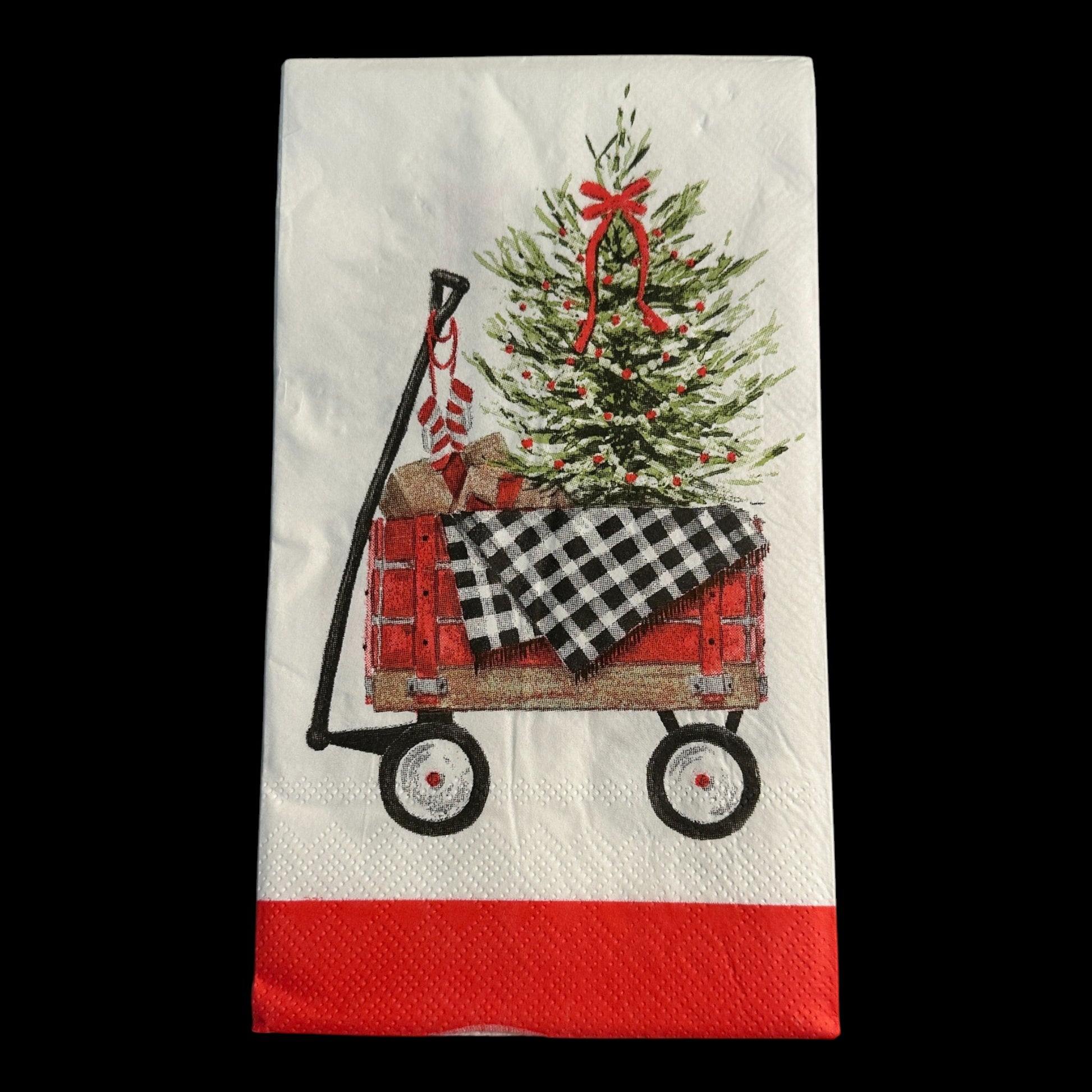 Close-up of a guest-size Christmas napkin featuring a red wagon carrying a decorated Christmas tree, wrapped gifts, and a black-and-white buffalo check blanket on a white background with a red border.
